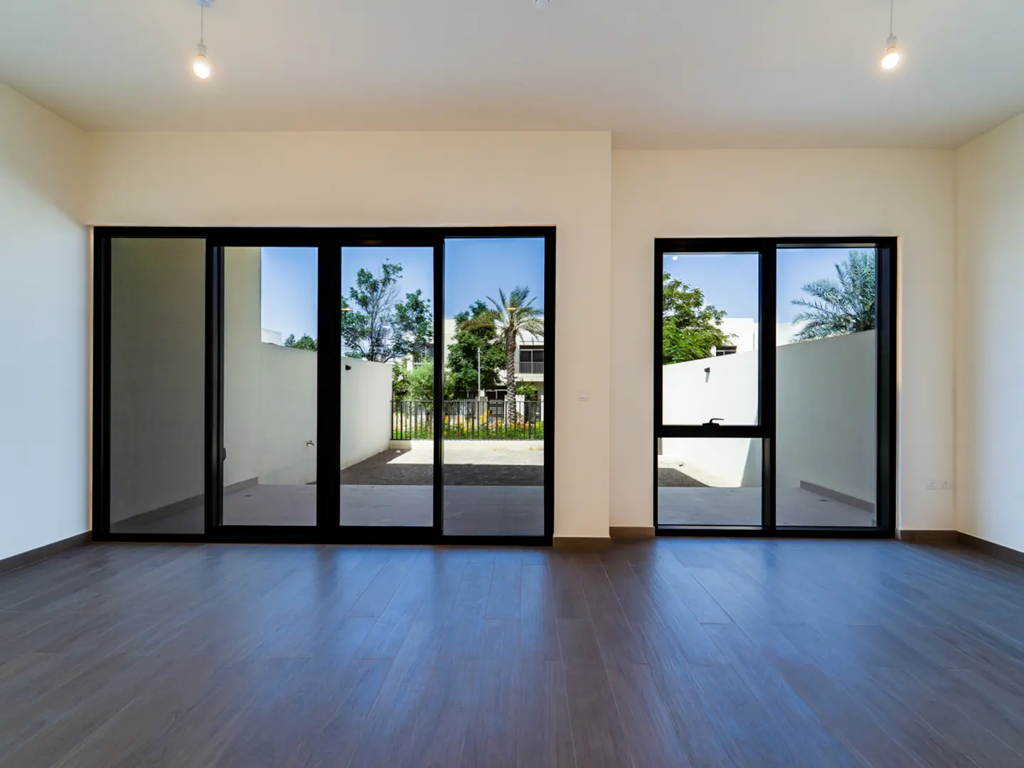 Empty room with dark wood floors, white walls, and black-framed sliding glass doors and windows showing a sunny outdoor area.