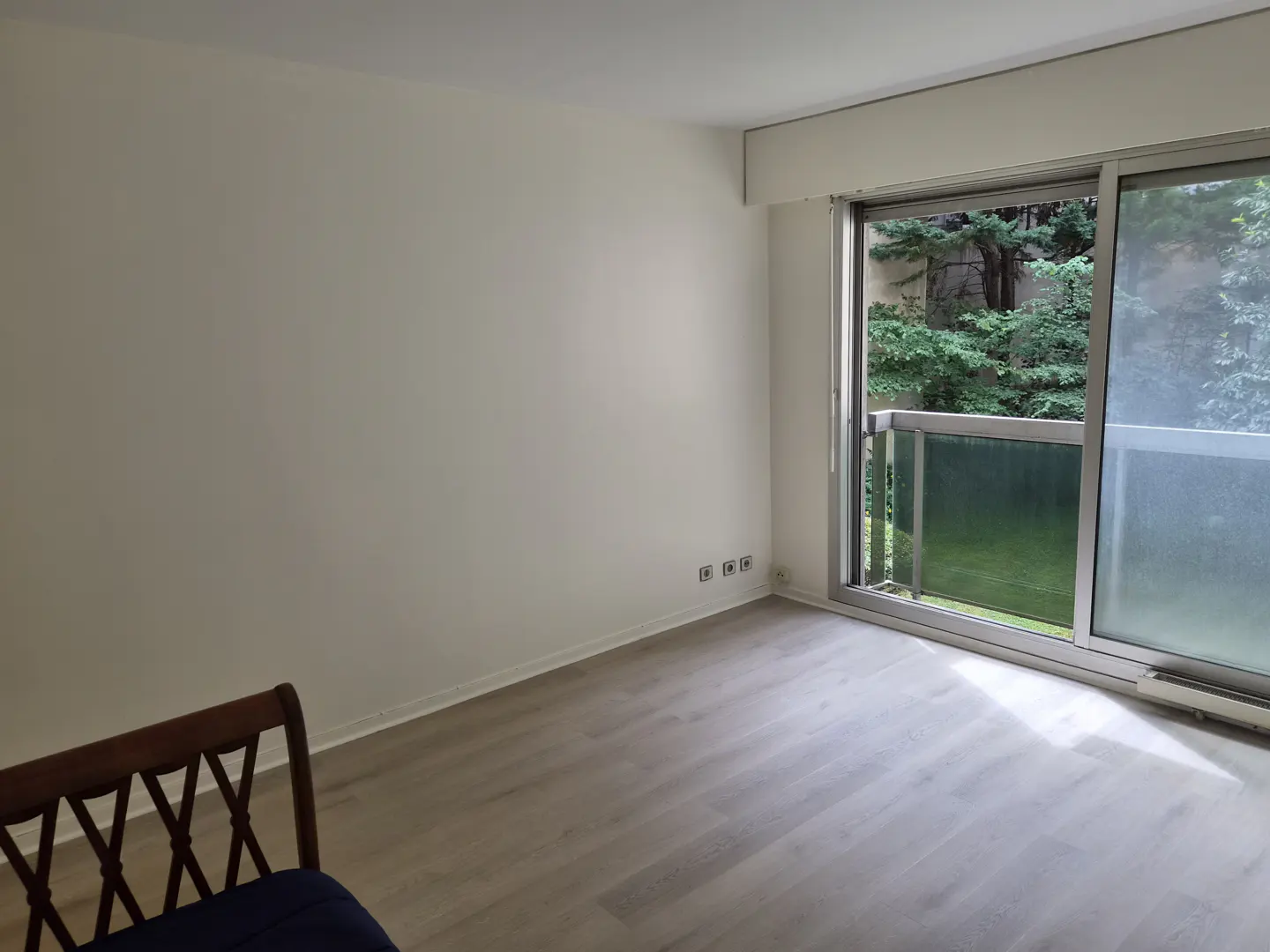 Empty room with light wood floors, white walls, and a sliding glass door leading to a balcony with green trees.