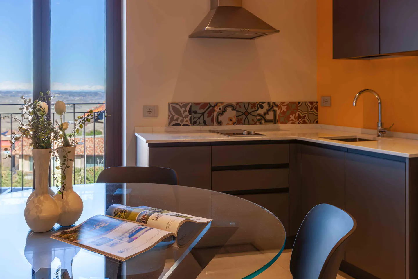 A modern kitchen with a glass table, vases, and an open magazine. The cabinets are gray, and the backsplash has colorful tiles. A window offers a scenic view.