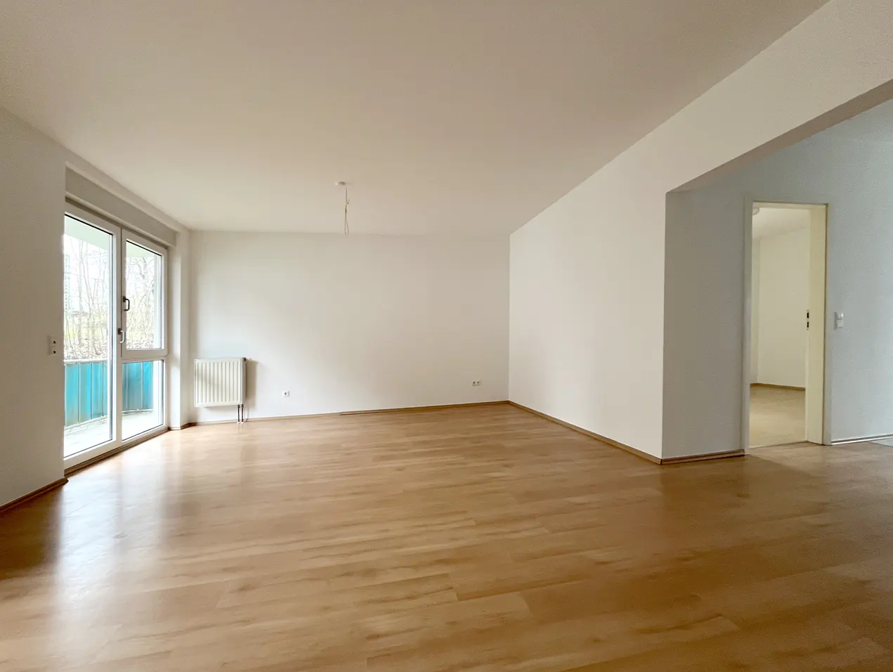 Bright, empty room with light wood floors, white walls, and a sliding glass door to a balcony. A doorway leads to another room.