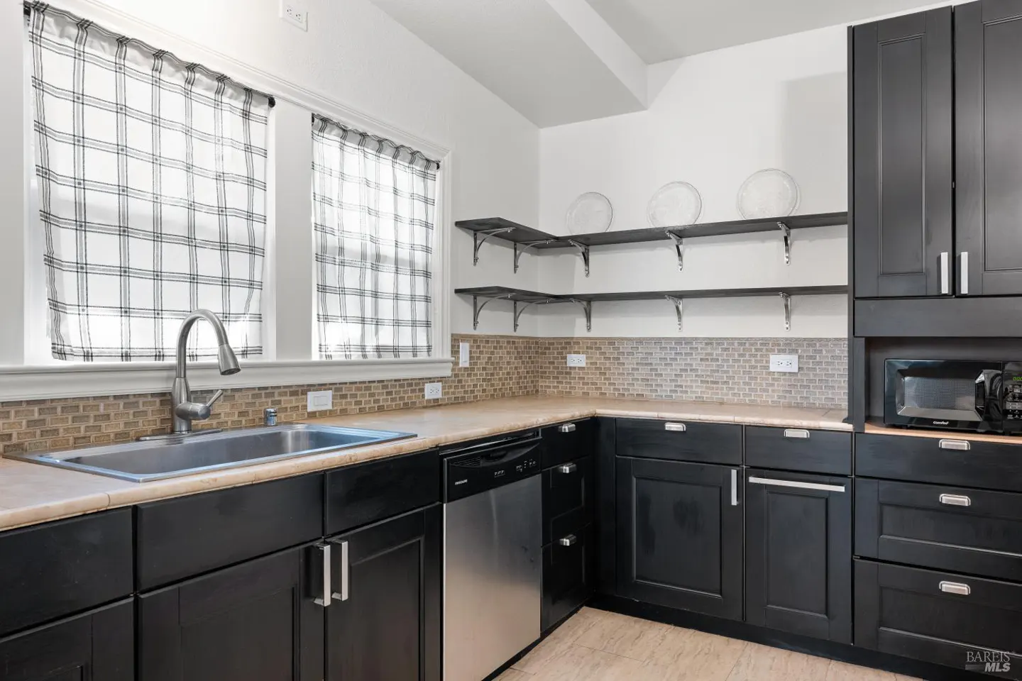 A kitchen with black cabinets, stainless steel appliances, and a sink under a window with plaid curtains.
