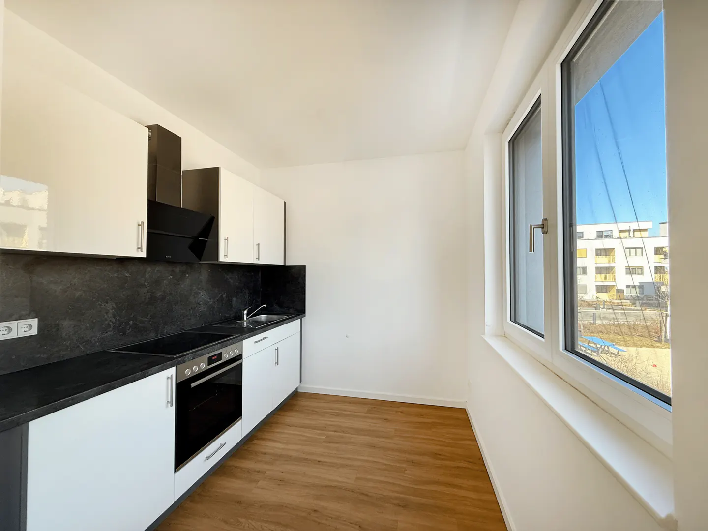 Modern kitchen with white cabinets, black countertops, built-in oven, and a large window overlooking a sunny neighborhood.
