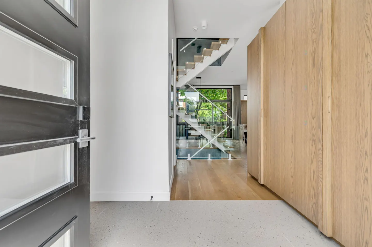 Modern entryway with a dark door, light wood cabinets, and a view of a glass-railed staircase leading to a bright living area.