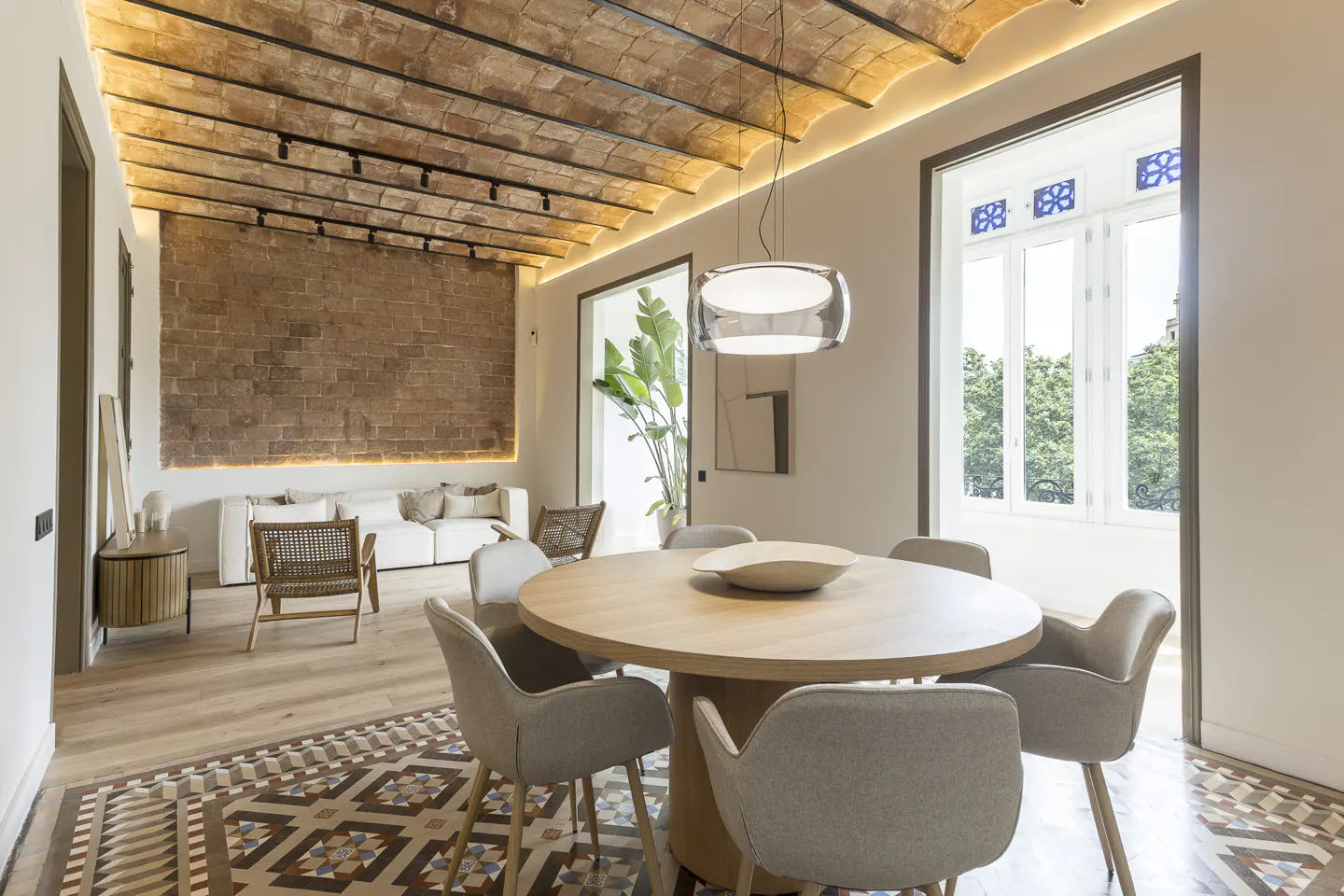 Modern dining room with a round wooden table, grey chairs, vaulted brick ceiling, and patterned tile flooring.