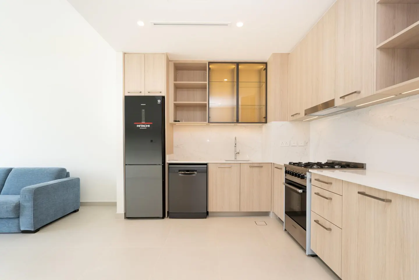 A modern kitchen with light wood cabinets, stainless steel appliances, and a blue sofa in the background.