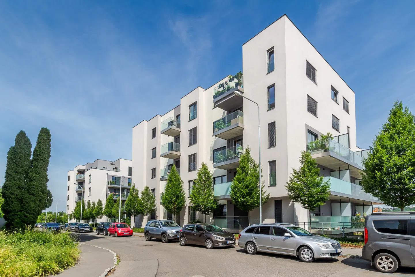 Modern white apartment buildings with balconies under a clear blue sky, with cars parked along a tree-lined street.