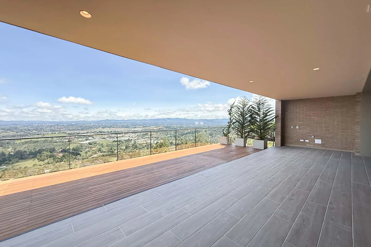A modern balcony with wood and tile flooring, glass railings, and potted trees overlooking a scenic landscape.