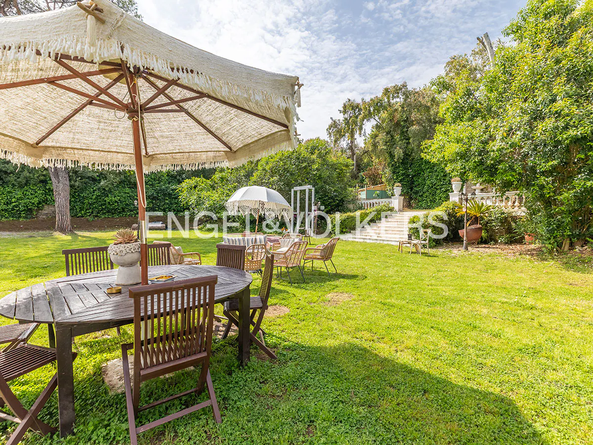 Outdoor patio with a wooden table and chairs under a fringed umbrella, set on a green lawn with trees and a white staircase in the background.