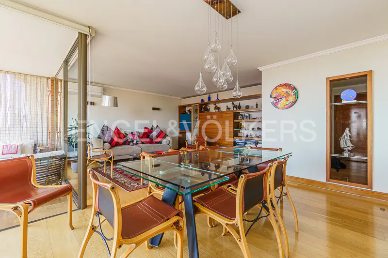Bright living room with a glass table, wooden chairs, and a beige sofa with colorful pillows. A unique chandelier hangs above the table.