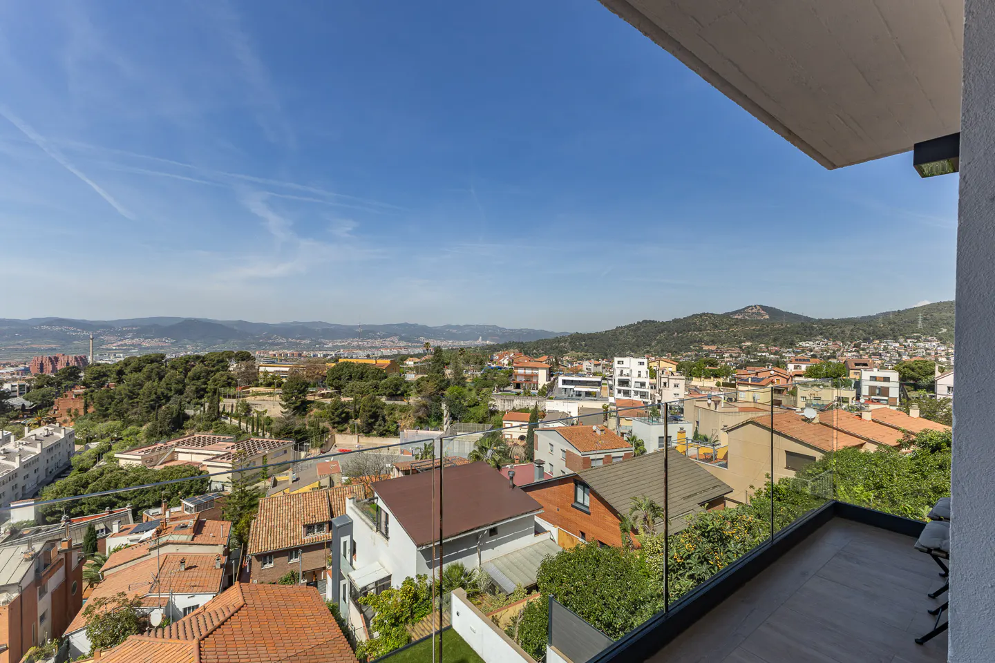 View from a modern balcony with a glass railing overlooking a suburban neighborhood with terracotta roofs and distant hills.