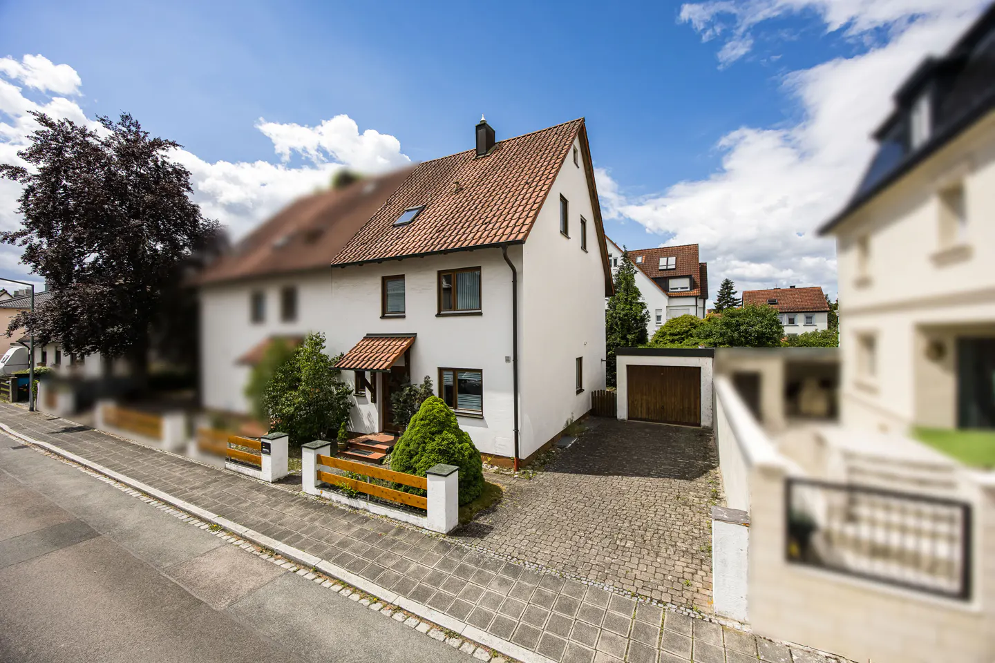 White two-story house with a red tile roof, a cobblestone driveway, a garage, and a small front garden under a blue sky.