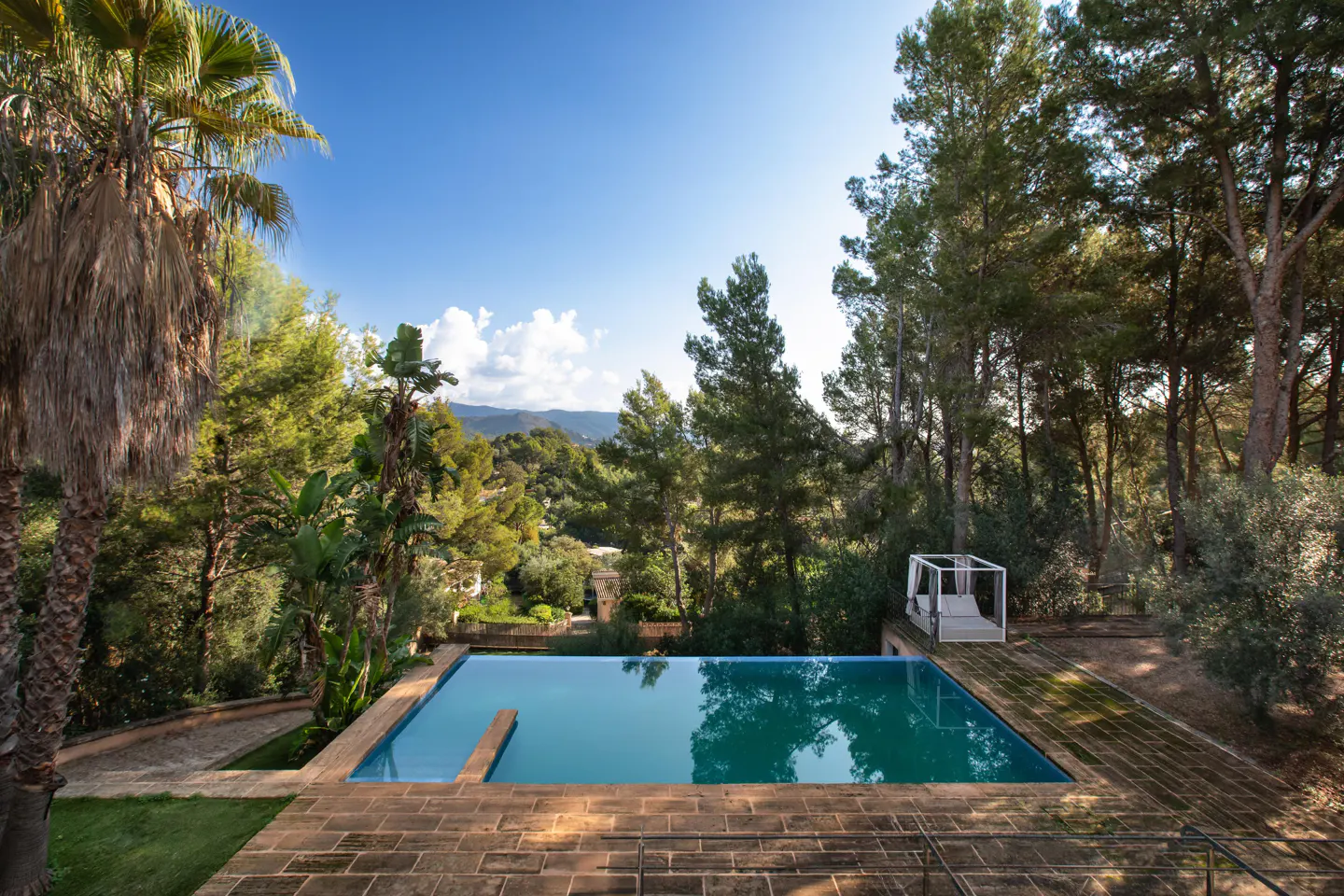 Outdoor pool with turquoise water surrounded by brick patio and lush green trees under a blue sky. A white cabana is visible.