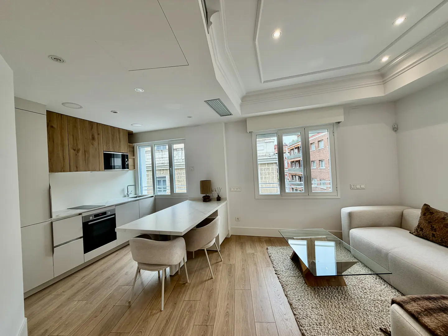 Bright, modern apartment with white walls, wood floors, and large windows. Kitchen with white cabinets and island, living area with sofa and glass table.
