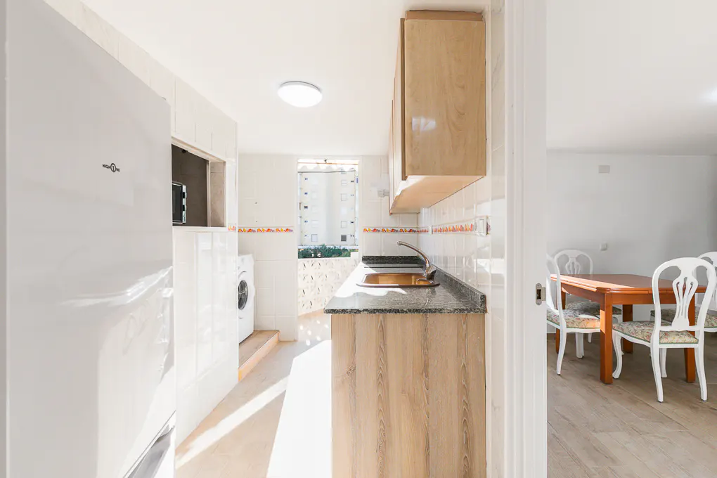Bright kitchen with white fridge, wood cabinets, and granite countertop. A dining room with a wood table and white chairs is visible.