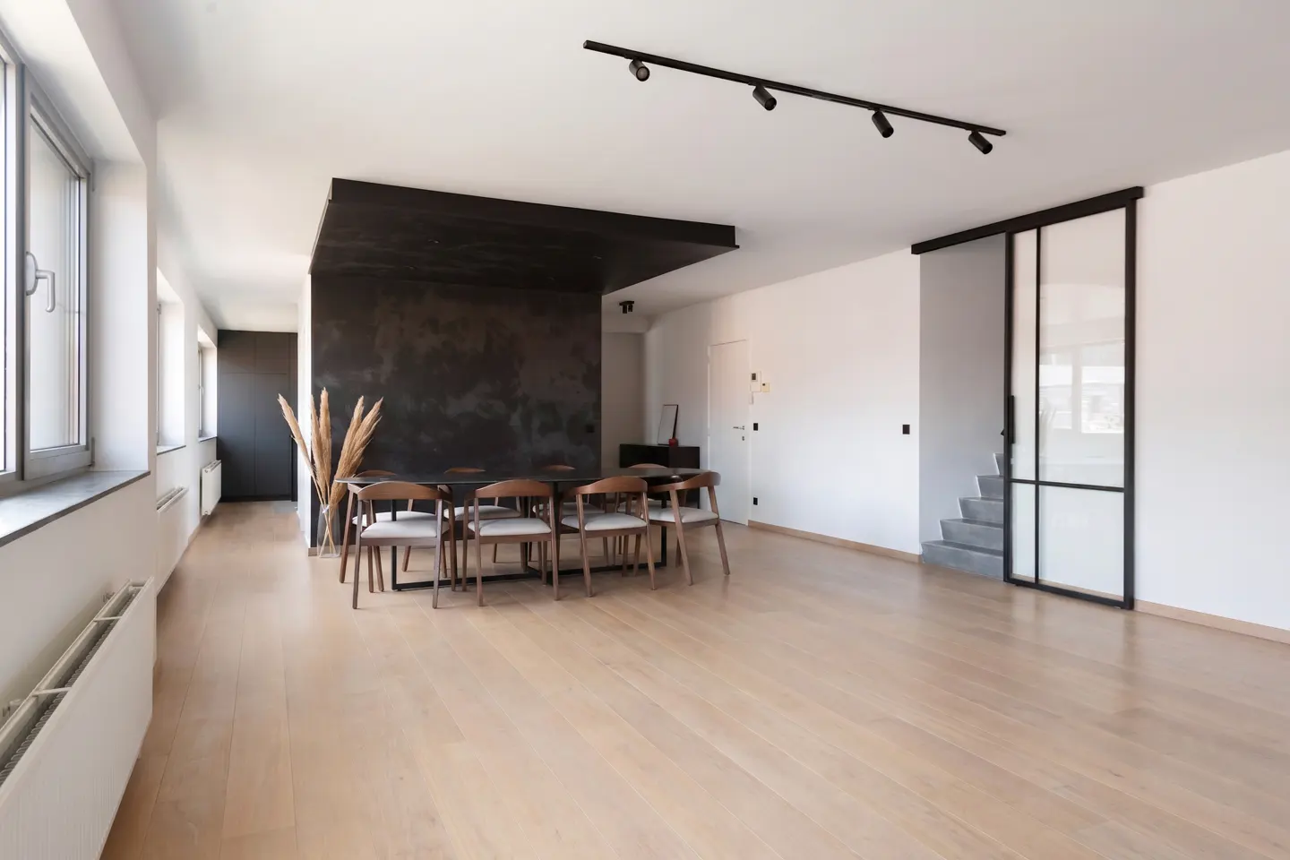 Bright, modern dining room with wood floors, a black accent wall, and a long table with chairs. Large windows line one wall.