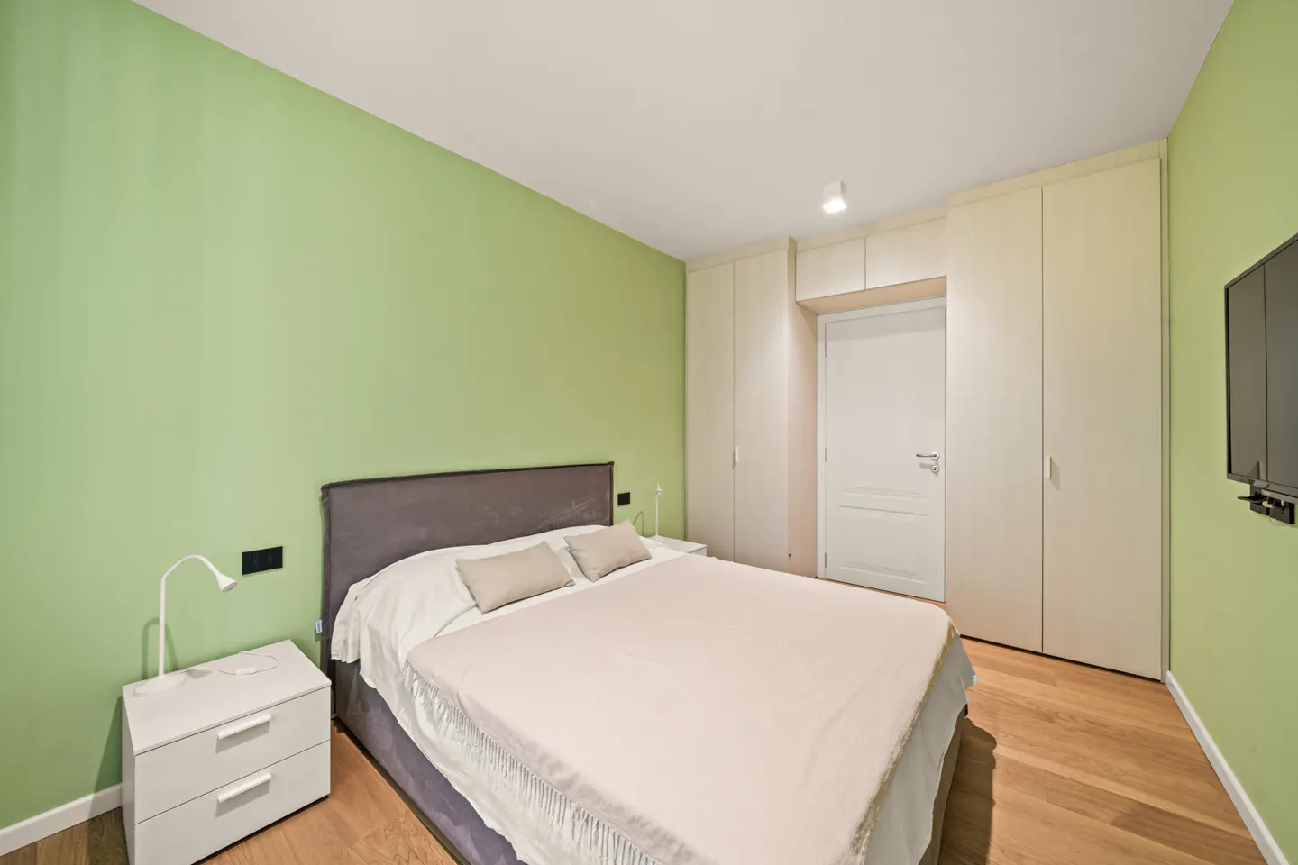 A bedroom with light green walls, a bed with a gray headboard, and a white nightstand. A white closet and door are in the background.