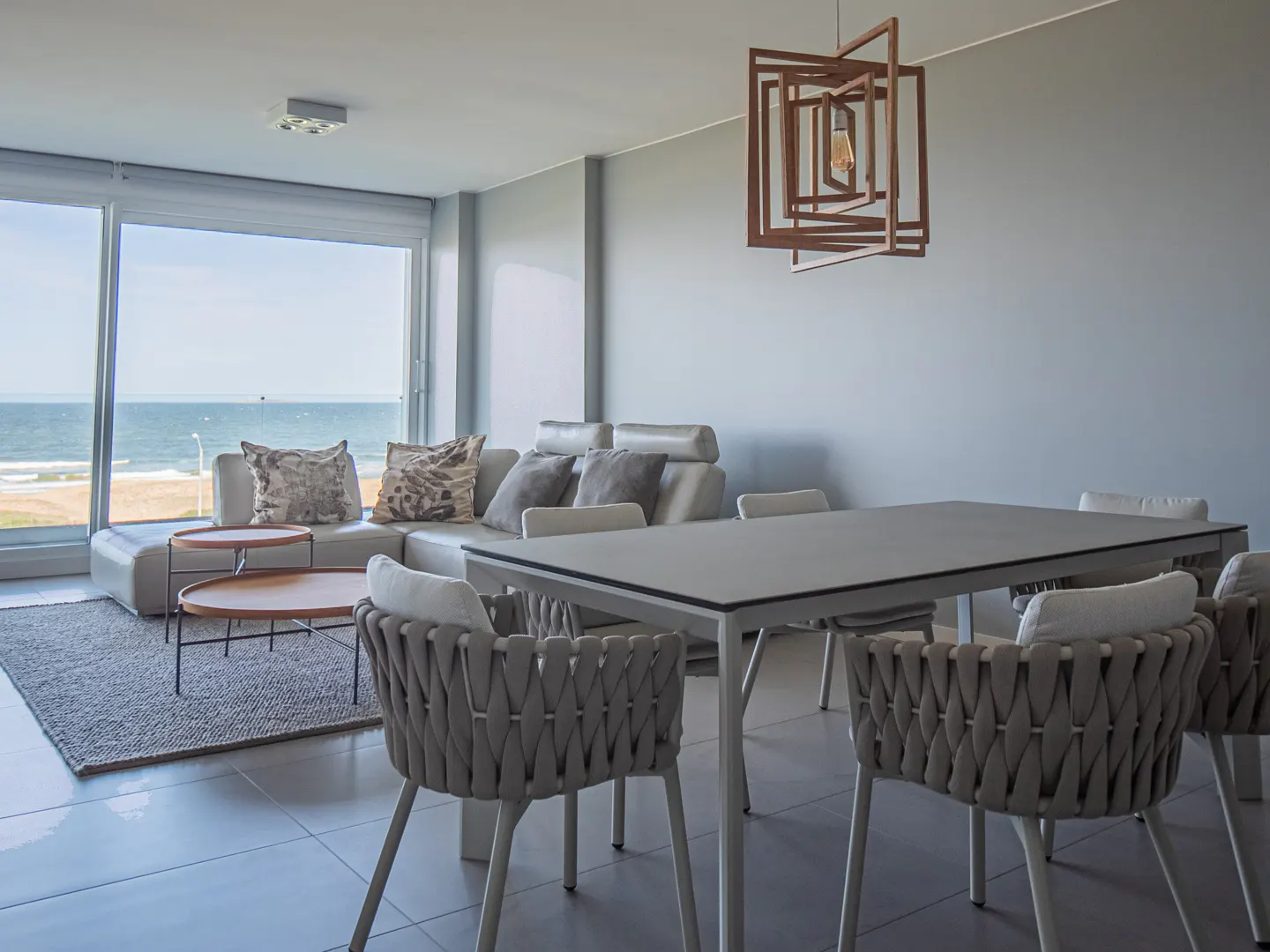 Living room with ocean view. Gray sofa with pillows, round tables, dining table with woven chairs, and a modern wooden light fixture.