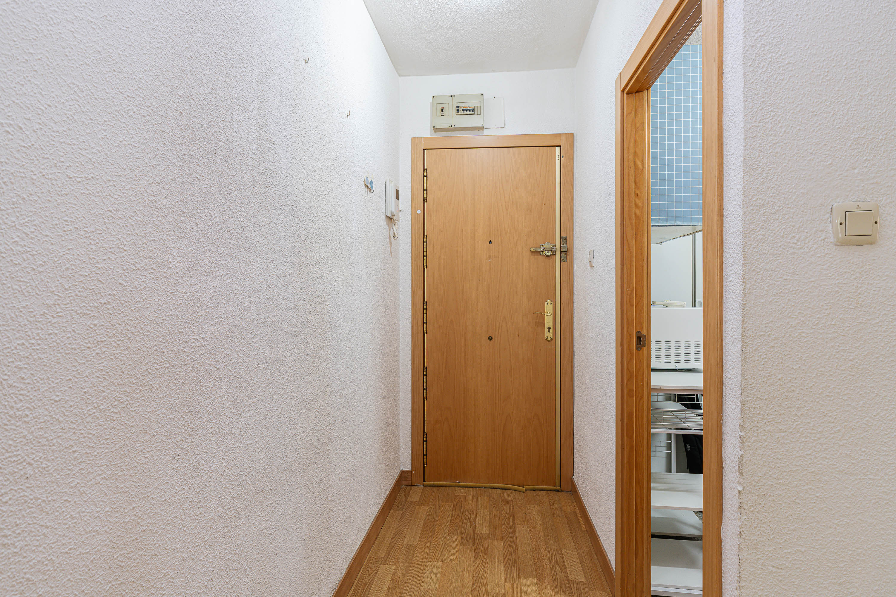 A narrow hallway with white walls, wood-look flooring, and a light wood front door, with a doorway leading to a kitchen area. [AI-generated text]