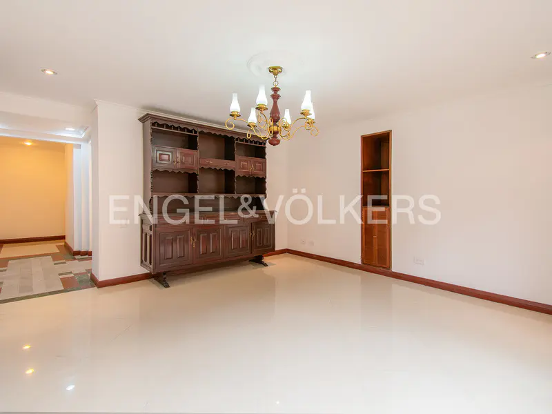 A bright, empty room with white walls and a dark wood hutch and chandelier.