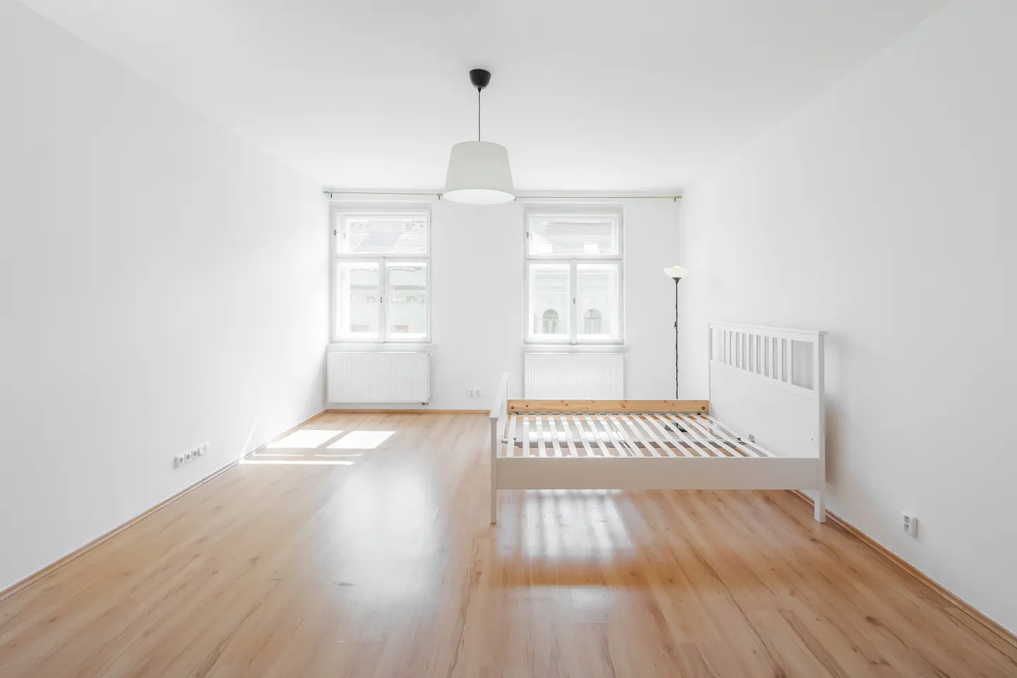 Bright, empty room with wood floors, white walls, two windows, and a white bed frame. A white pendant light hangs from the ceiling.