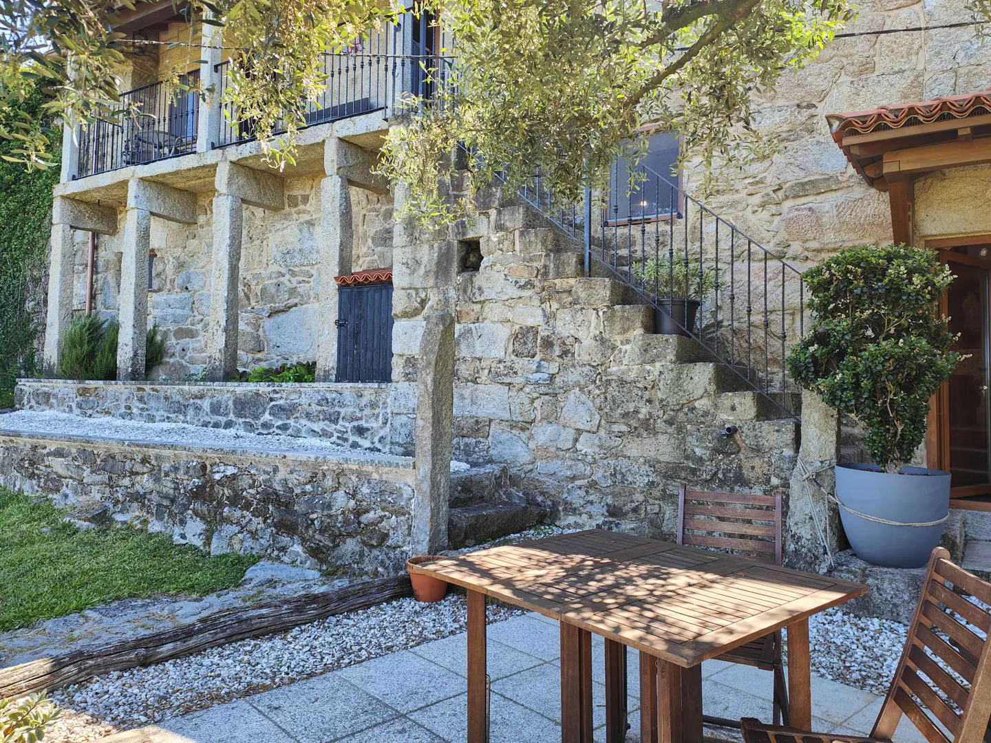 Rustic stone house with a balcony, outdoor stone stairs, and a wooden patio table set on a paved terrace.