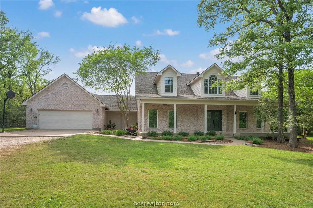 Two-story brick house with a covered porch, dormer windows, and a two-car garage, surrounded by green lawn and trees.