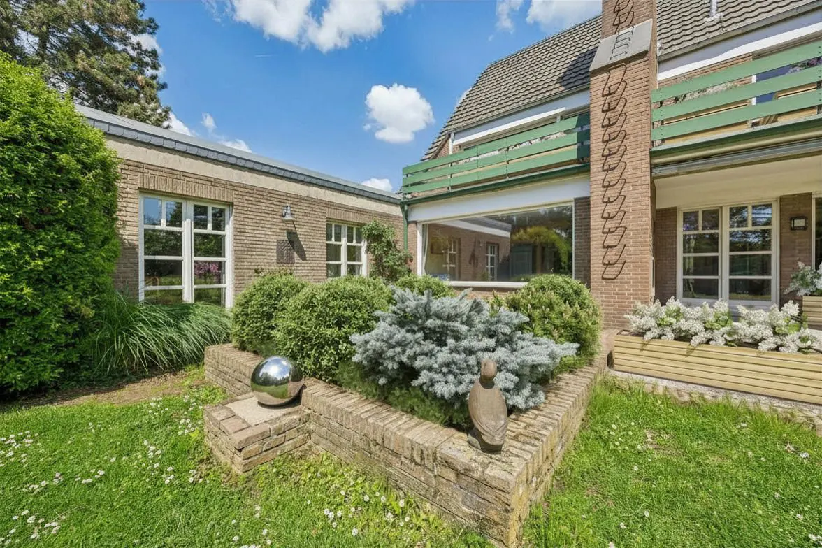 Exterior view of a brick house with a green balcony, manicured bushes, and a green lawn.