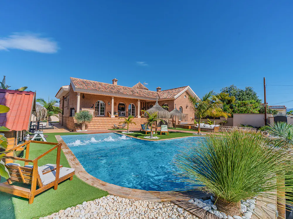A brick house with a red tile roof overlooks a blue pool with lounge chairs and palm trees under a clear blue sky.