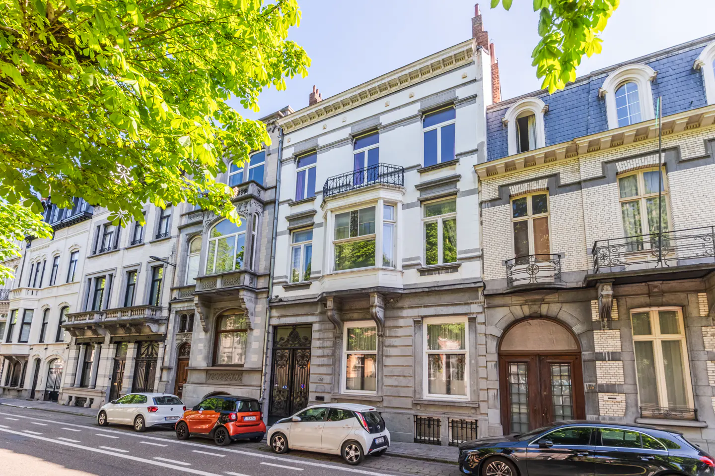 Street view of European-style townhouses with parked cars. Buildings are white and gray with blue accents. Green trees are in the foreground.