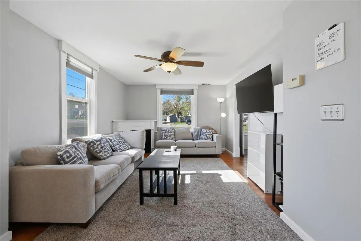 Living room with gray walls, a ceiling fan, and two sofas with patterned pillows. A black coffee table sits on a gray carpet. A TV is mounted on the wall.