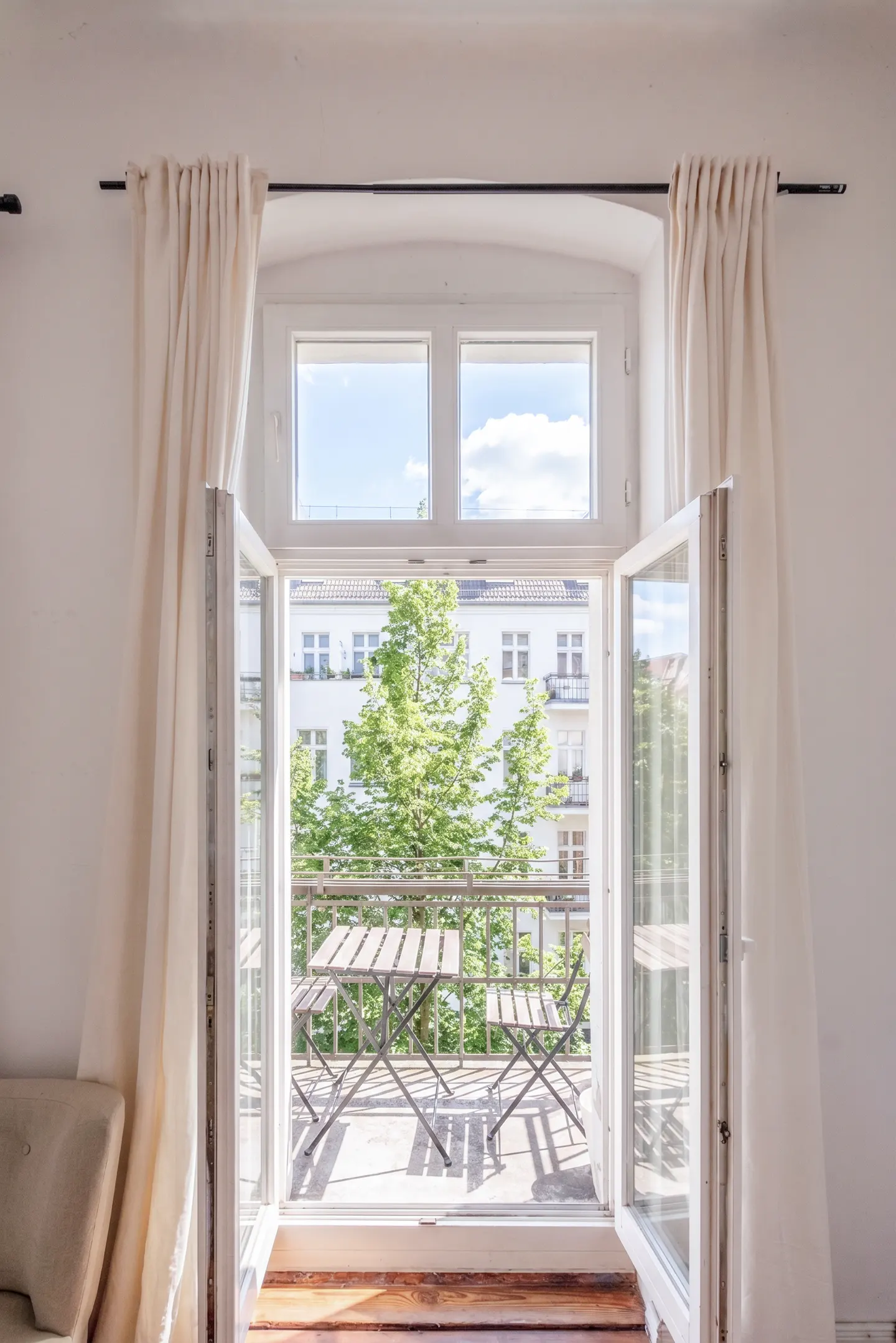 Open white French doors lead to a balcony with a table and chairs, offering a view of a tree and building. Cream-colored curtains frame the doorway.