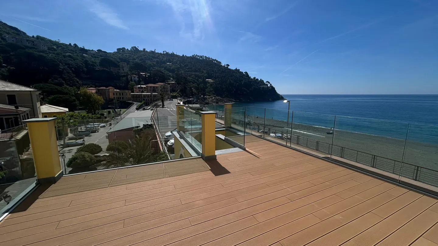 A wood-look deck with glass railings overlooks a beach and blue ocean on a sunny day. Green hills rise in the background.