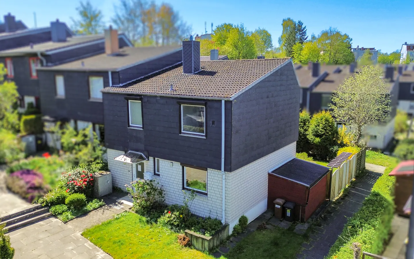 Two-story townhouse with a dark gray facade, white brick base, and a brown roof. Green lawn and garden in front. Blue sky.