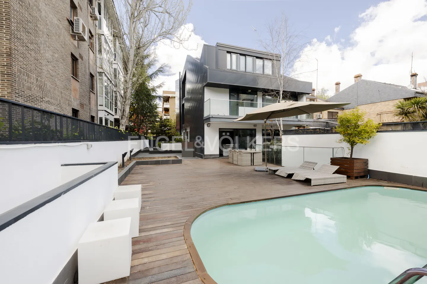 Modern home exterior with a wooden deck, swimming pool, lounge chairs, and a large umbrella under a bright sky.