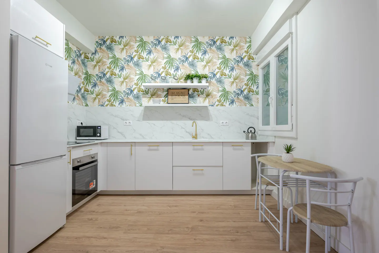 Bright kitchen with white cabinets, gold hardware, and tropical wallpaper. A small table and chairs sit near a window.
