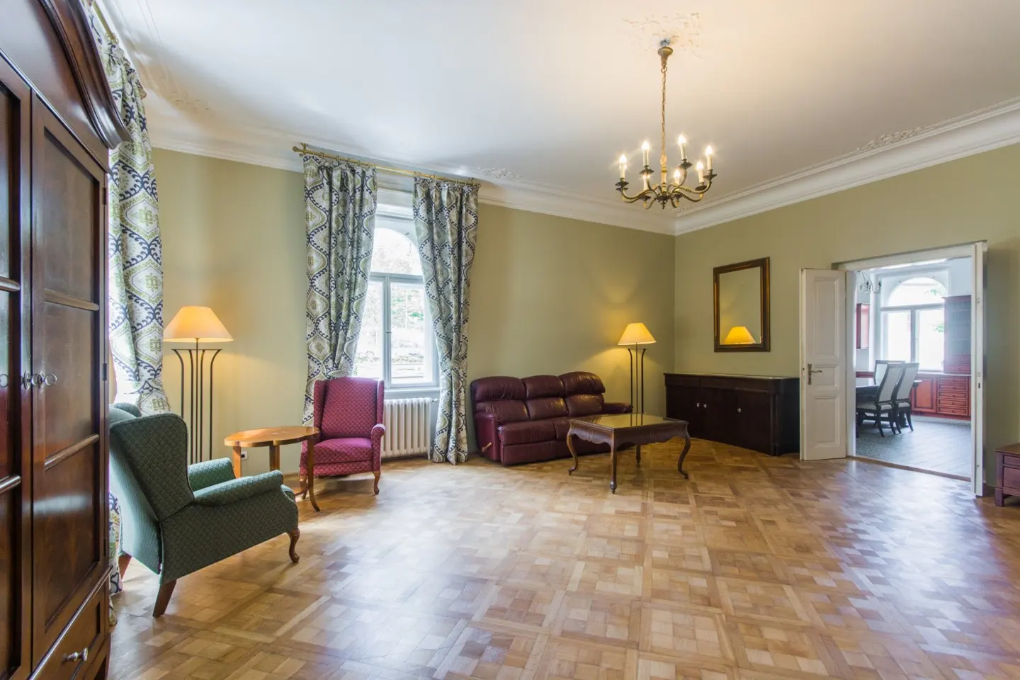 A living room with parquet floors, olive green walls, and a chandelier. There is a brown leather sofa, two armchairs, and a dark wood cabinet.