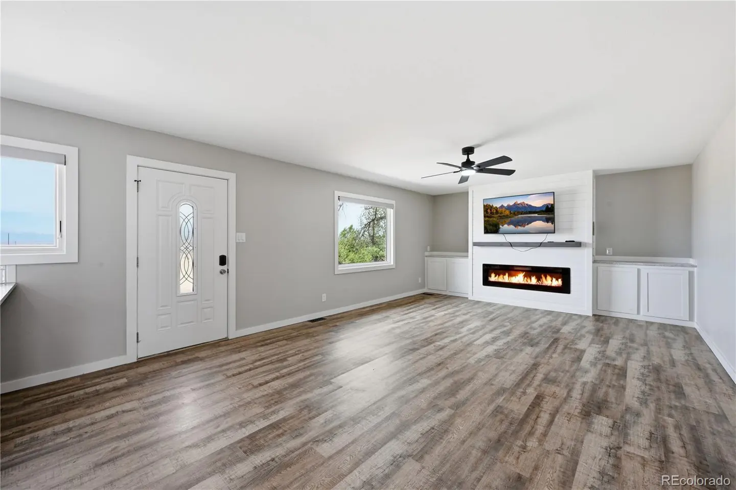 Bright living room with wood-look floors, white walls, a modern fireplace, wall-mounted TV, and a white front door.