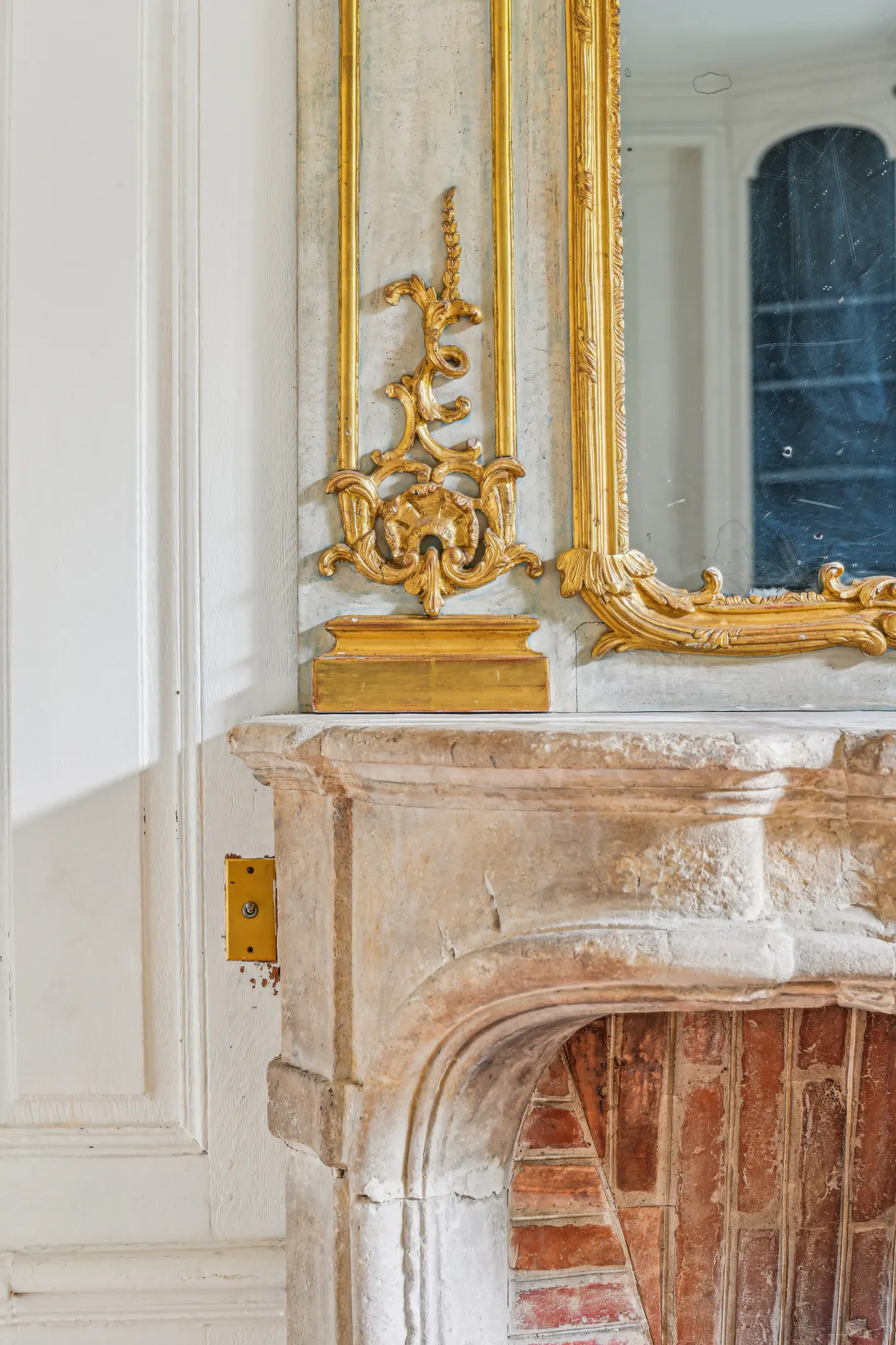 Ornate gold mirror frame resting on a rustic stone fireplace mantel with exposed red brick interior.