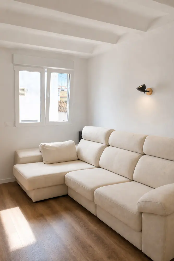 Bright living room with a large, beige sectional sofa, wood floors, white walls, and a window letting in natural light.