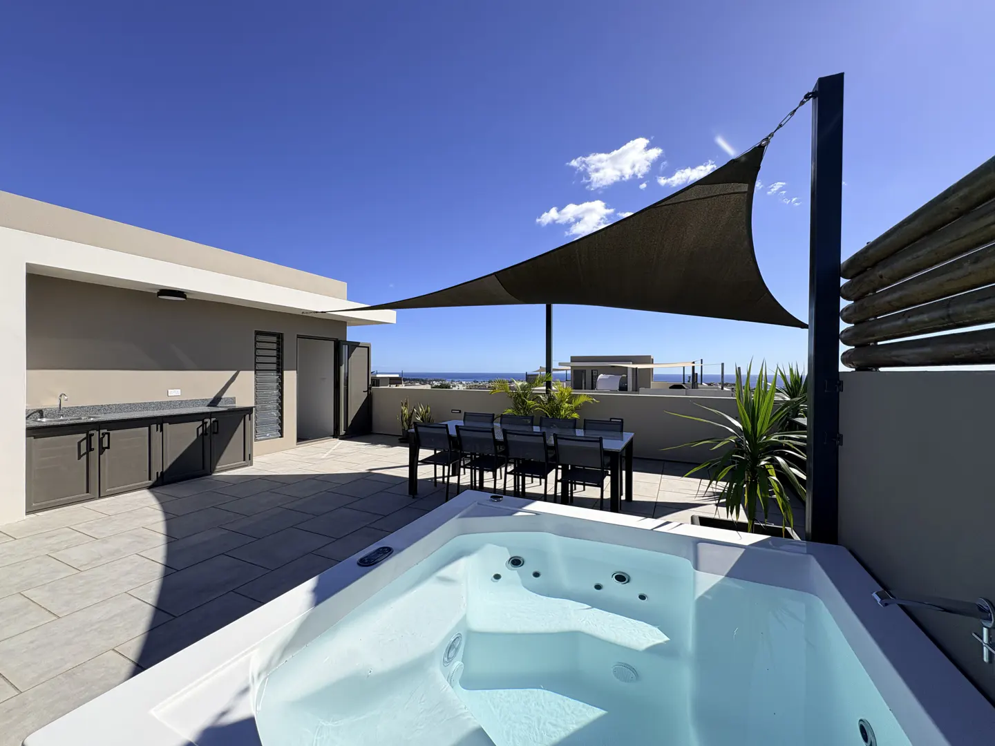 Rooftop patio with a white hot tub, dining table, and outdoor kitchen under a black sunshade against a blue sky.