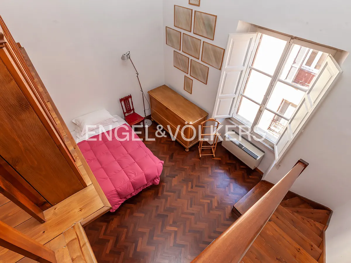 A high-angle shot of a bedroom with a red bedspread, wooden floors, and a large window with white shutters.