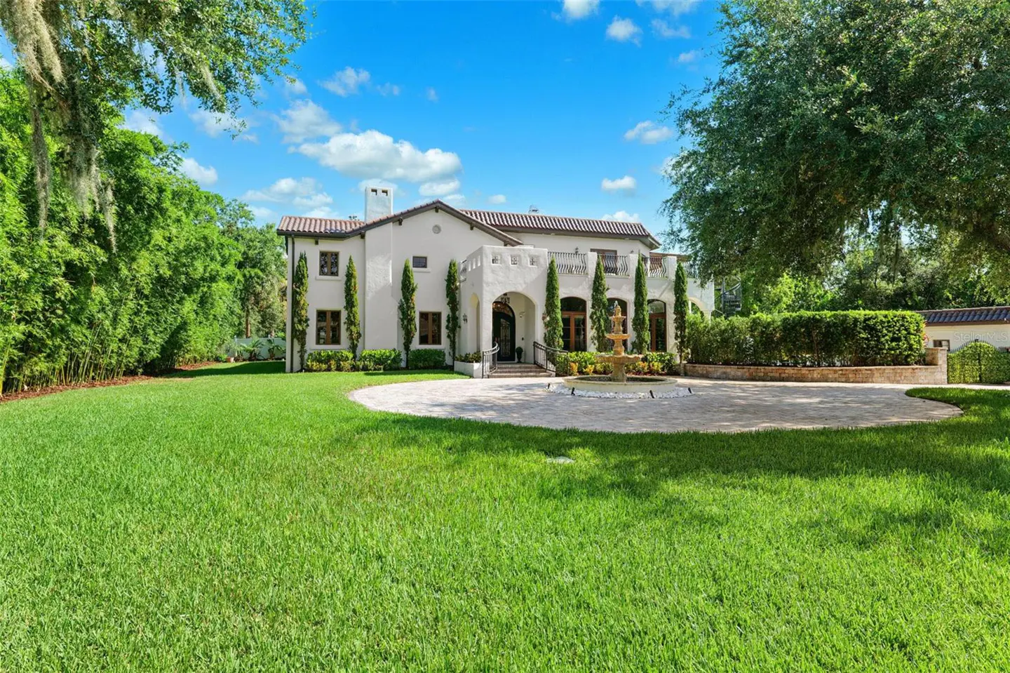A two-story white house with a red tile roof, a fountain, and a green lawn on a sunny day.