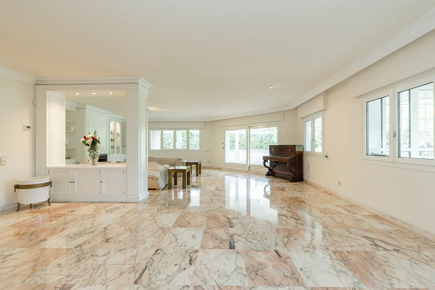 Bright, spacious living room with marble floors, white walls, and large windows. A piano sits near the windows, and a white cabinet with flowers is on the left.