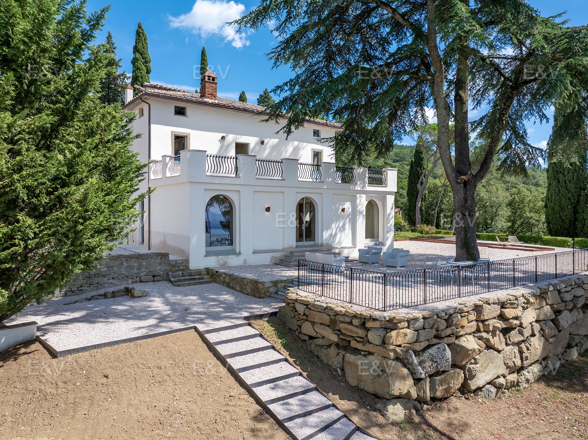 White villa with a terrace, stone retaining wall, and gravel patio under a large tree on a sunny day. [AI-generated text]