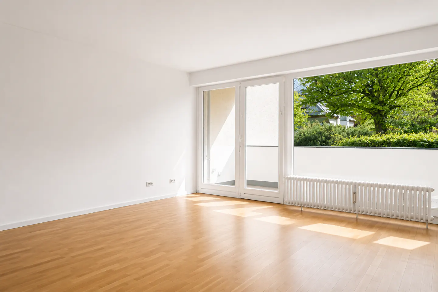 Empty room with light wood floors, white walls, a radiator, and glass doors opening to a balcony with green trees.
