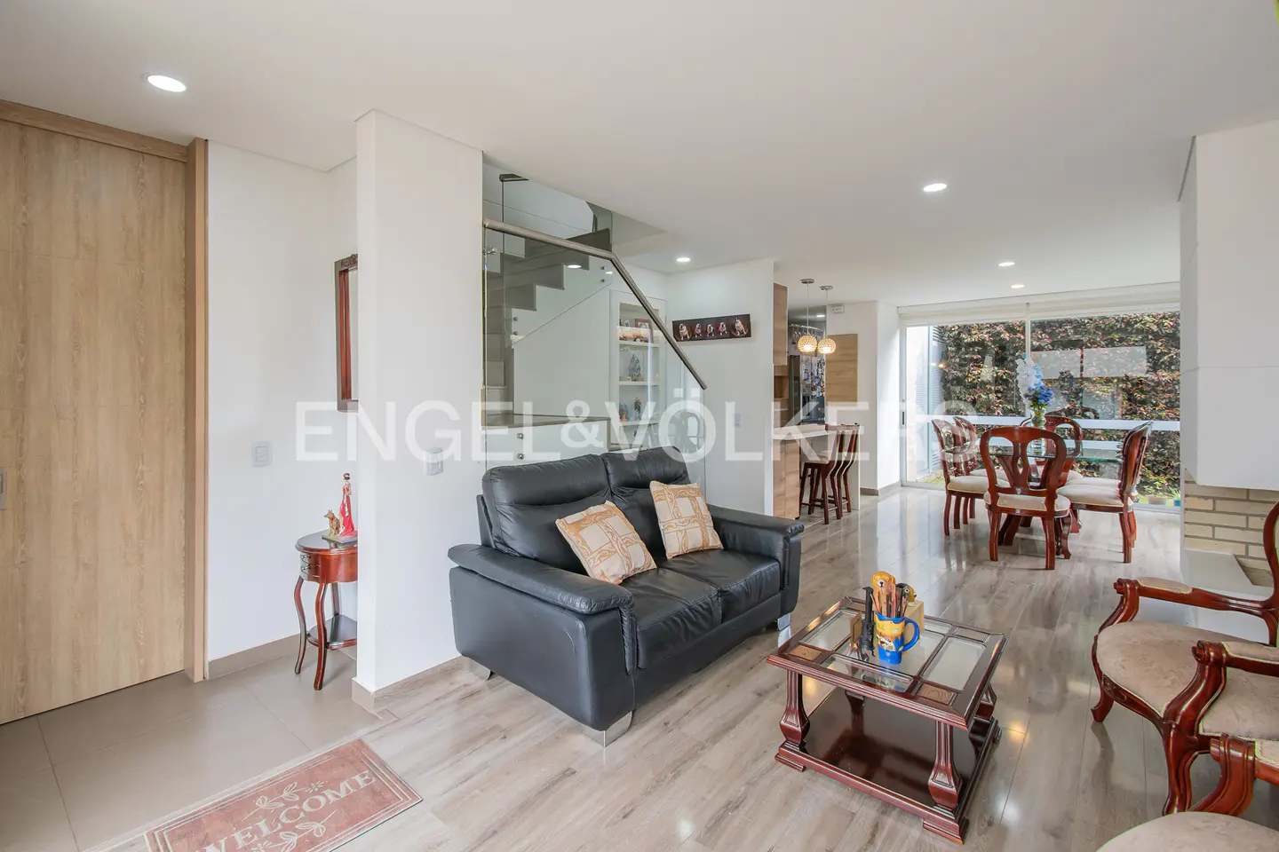 Modern living room with a black leather sofa, glass coffee table, and dining area with wooden chairs and large windows.