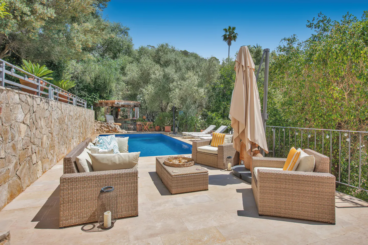Outdoor patio with wicker furniture, a pool, and lush greenery under a clear blue sky.