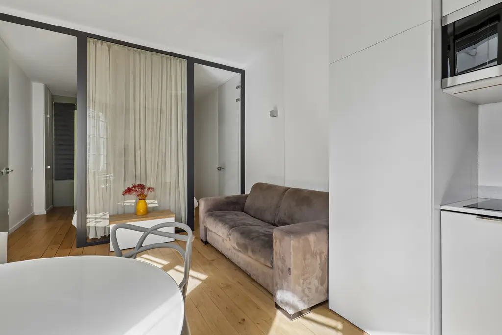 Living room with a brown sofa, white walls, and wood floors. A glass partition separates the living area from another room. A yellow vase with flowers sits on a table.