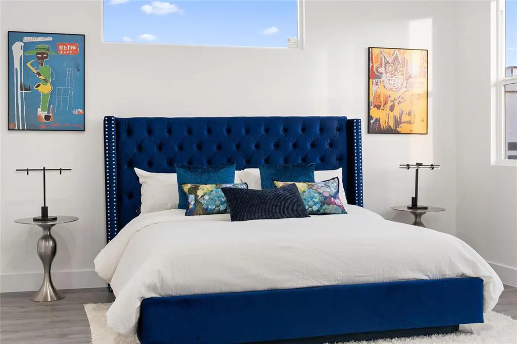 Bedroom with a blue velvet bed, white linens, and decorative pillows. Modern art hangs on the white walls. Side tables with lamps flank the bed.