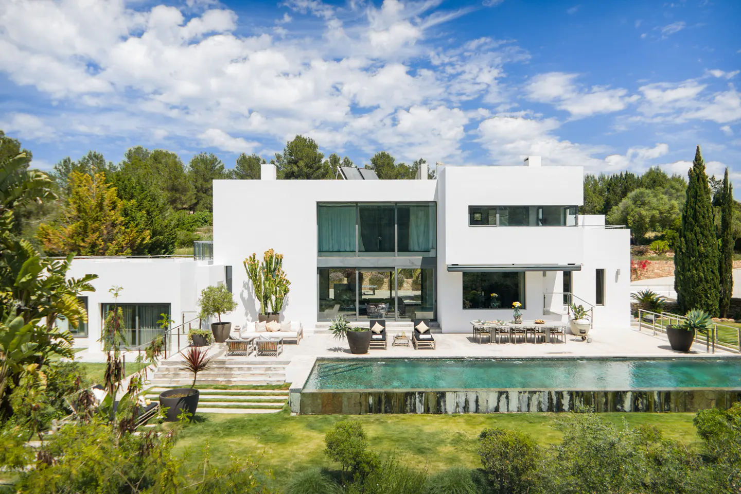 Modern white house with a pool, patio furniture, and green trees under a blue sky.